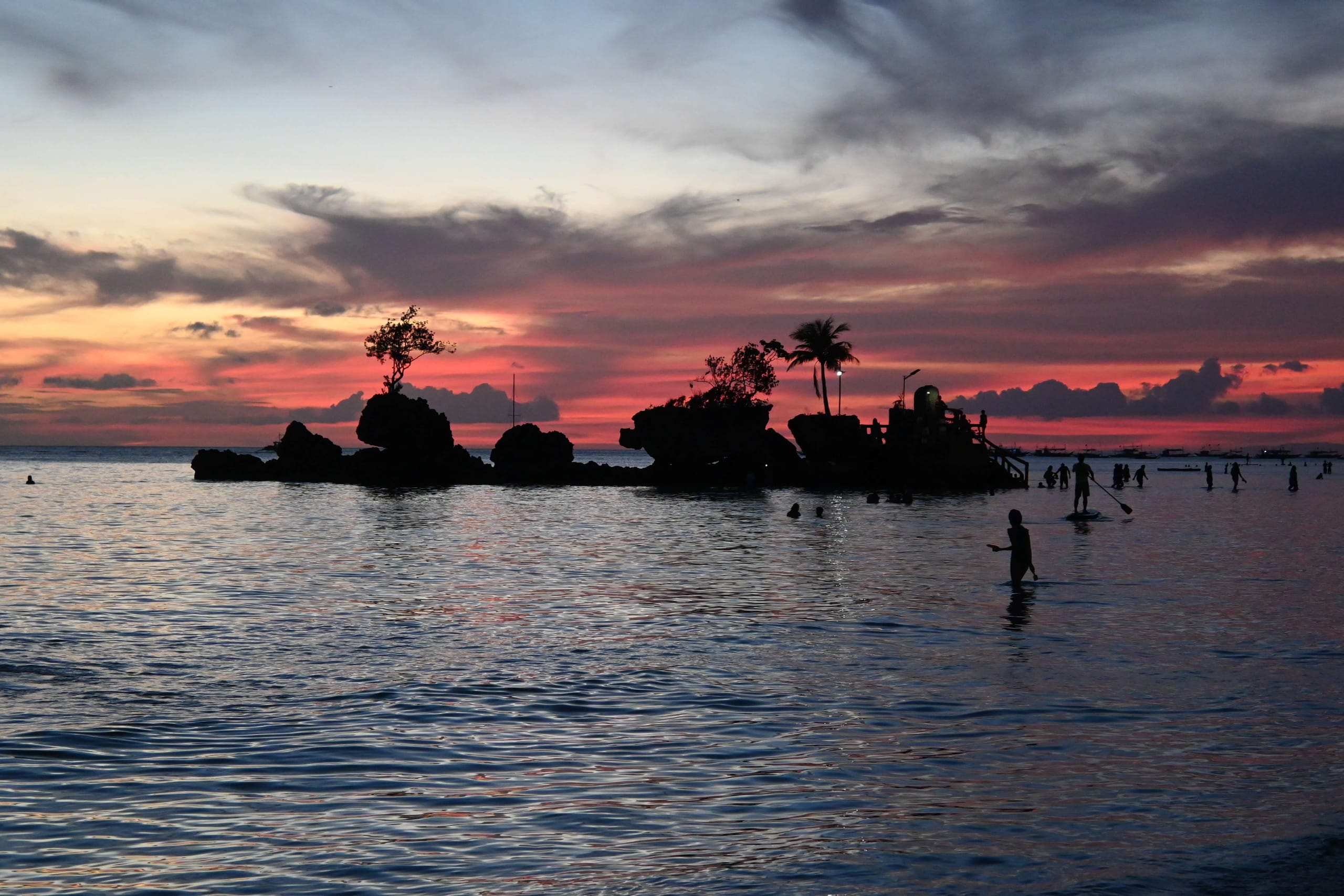 Willy's Rock off White Beach in Boracay, silhouetted at sunset with its shrine steps and a single palm against a red sky, people standing in the shallows