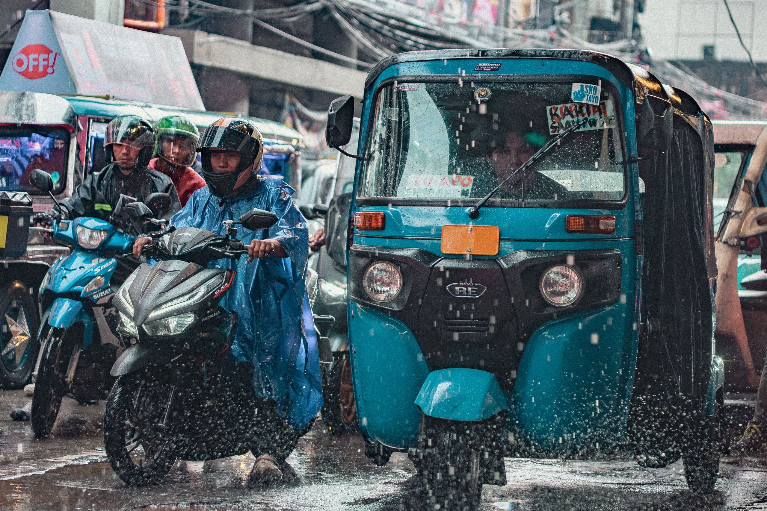 Motorbikes and a teal auto rickshaw stopped on a Manila street in heavy rain, one rider in a blue plastic poncho