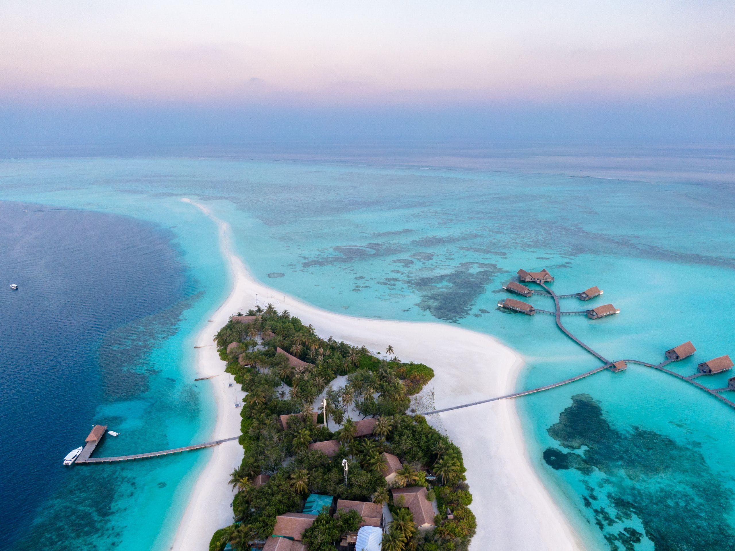 A resort island with a long white sand spit, a boat jetty and overwater villas, seen from above