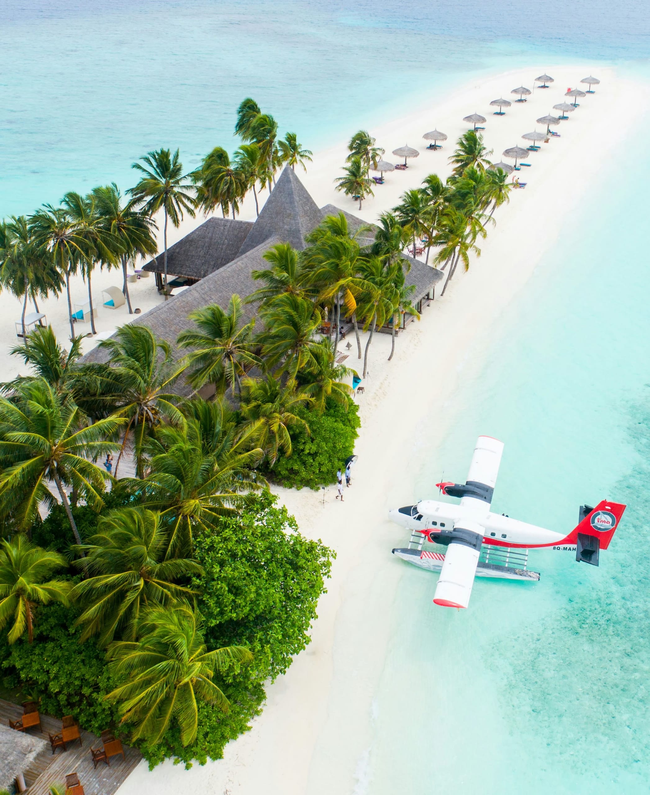 A red and white seaplane resting on the sand beside palms and beach umbrellas