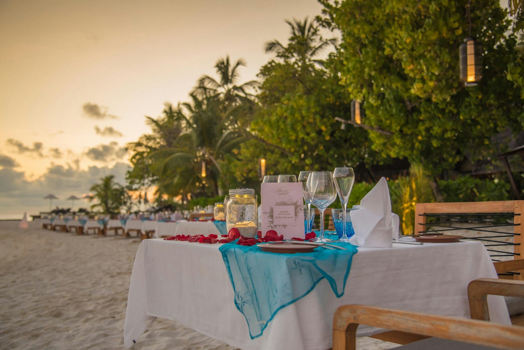 A long table laid for dinner on the sand at dusk, glasses and a lantern set out under the palms