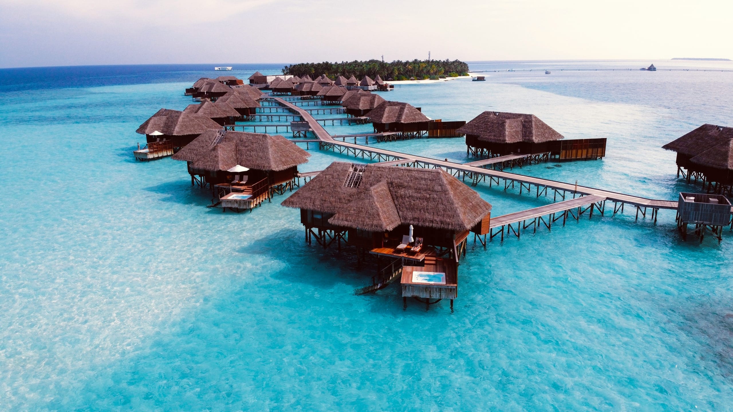 Thatched overwater villas on stilts linked by boardwalks over pale turquoise shallows, with the palm-covered island behind them
